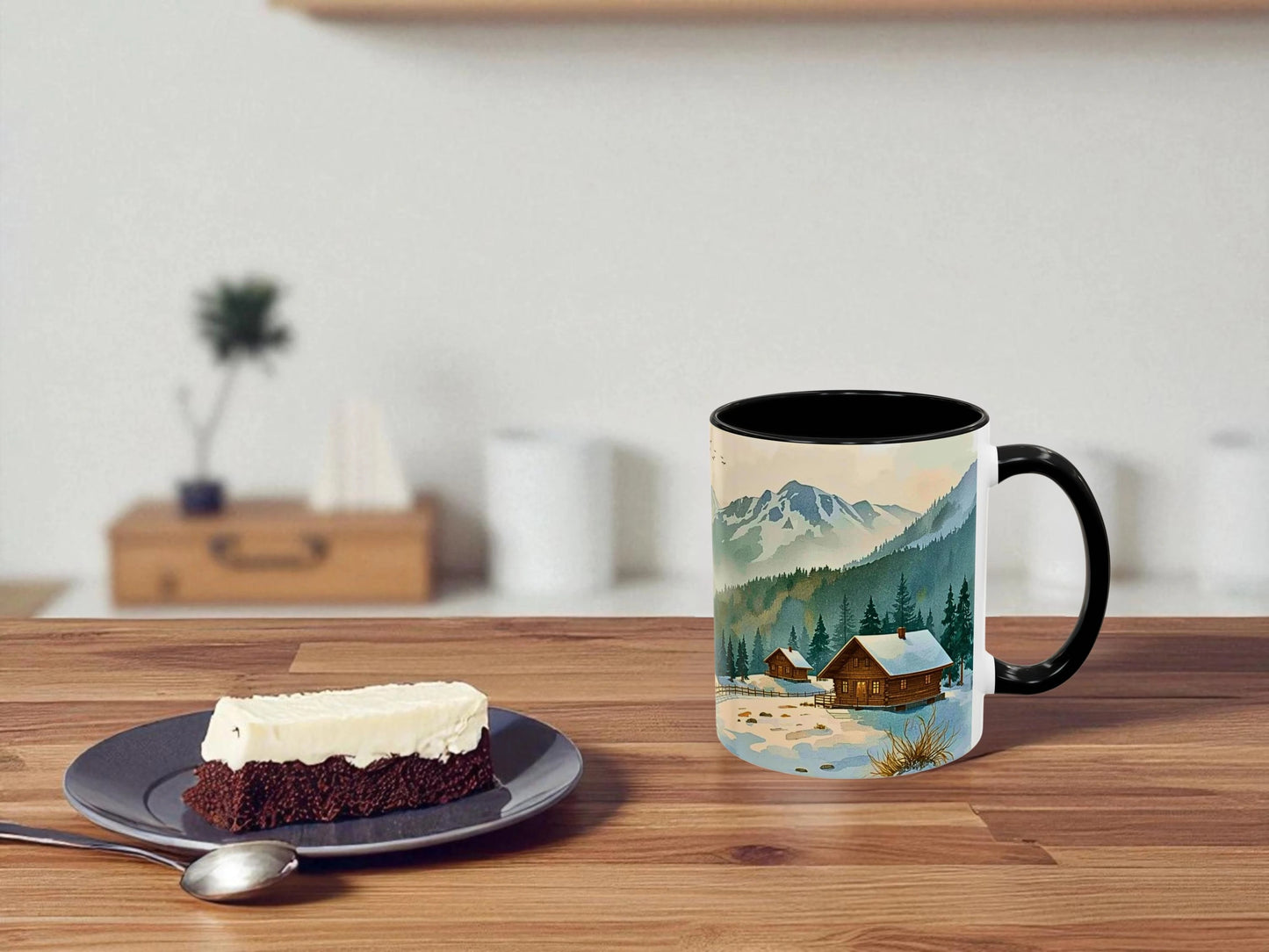 Mug with mountain and cabin design on a wooden table with a piece of cake.