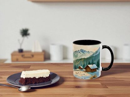 Mug with mountain and cabin design on a wooden table with a piece of cake.