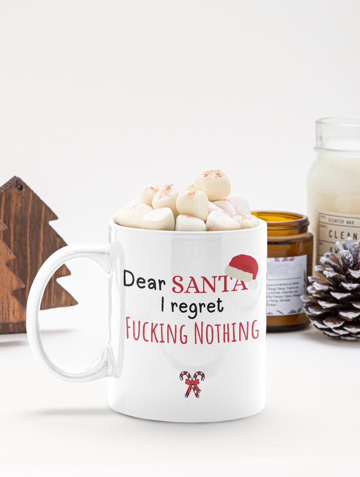 White mug with text 'Dear Santa I regret nothing' filled with hot chocolate and marshmallows, on a white surface with a candle and decorative items.