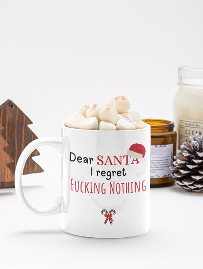 White mug with text 'Dear Santa I regret nothing' filled with hot chocolate and marshmallows, on a white surface with a candle and decorative items.