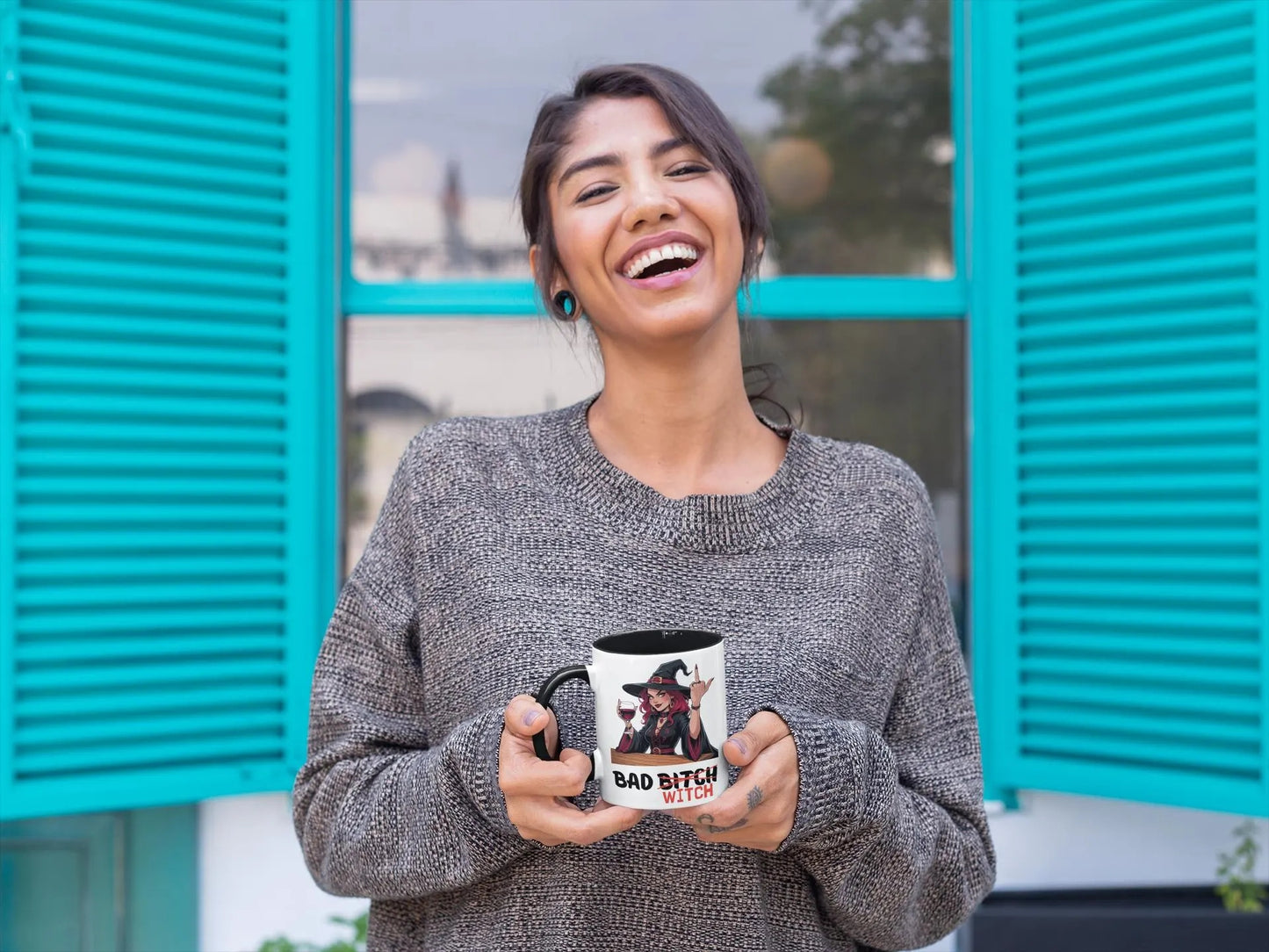 Woman holding a mug with text, standing in front of teal shutters.