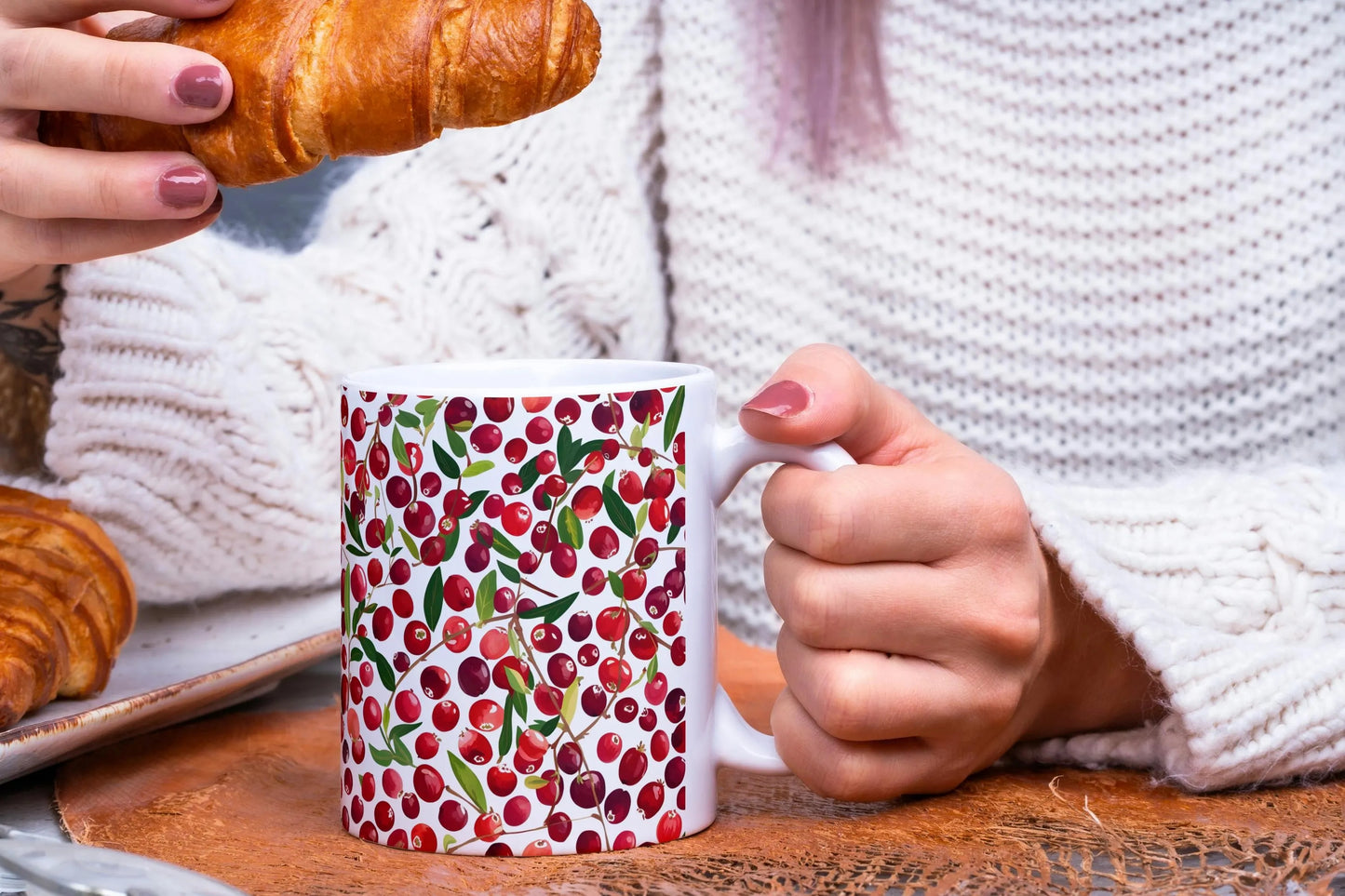 Person holding a mug with a cranberry pattern and a croissant on a wooden table.
