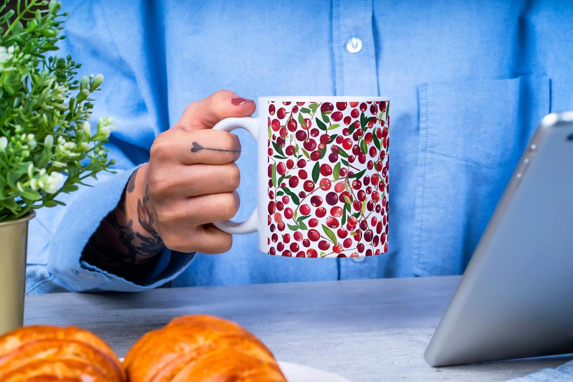 Person holding a mug with a cranberry pattern at a table with pastries and a laptop.