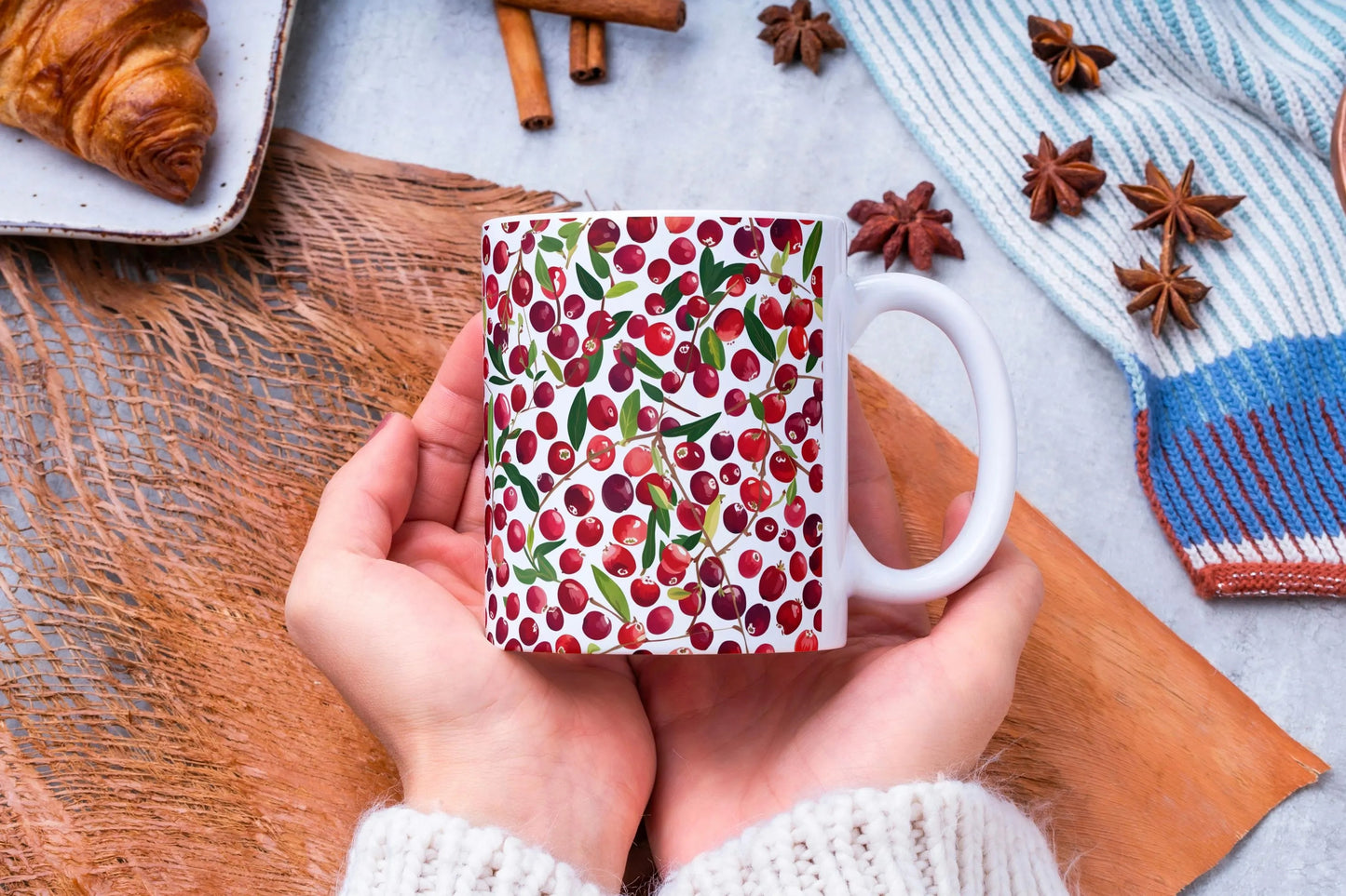 Hand holding a mug with a cranberry pattern on a wooden surface with cinnamon sticks and star anise.
