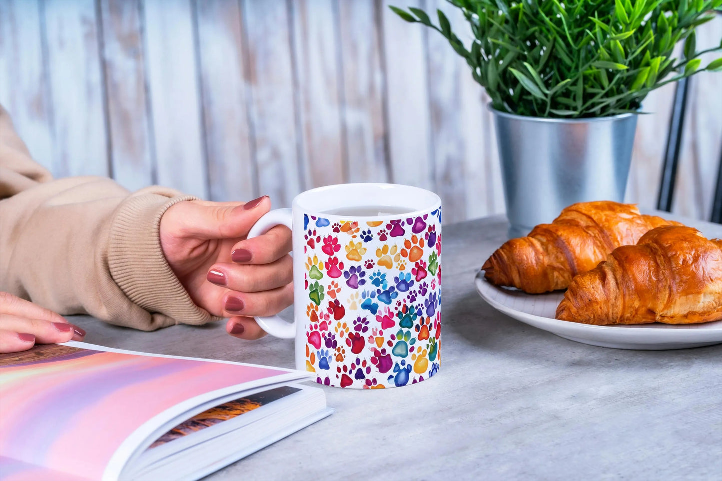 Person holding a mug with colorful paw prints, next to a plate of croissants and a plant on a table.
