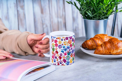 Person holding a mug with colorful paw prints, next to a plate of croissants and a plant on a table.