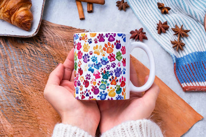 Hand holding a mug with colorful paw prints, surrounded by cinnamon sticks and a croissant on a wooden surface.
