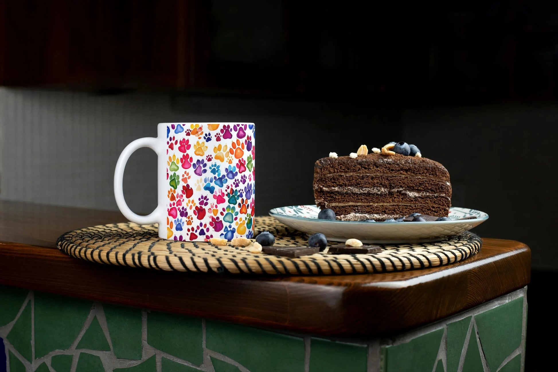 Mug with colorful paw print pattern next to a chocolate cake on a wooden surface.