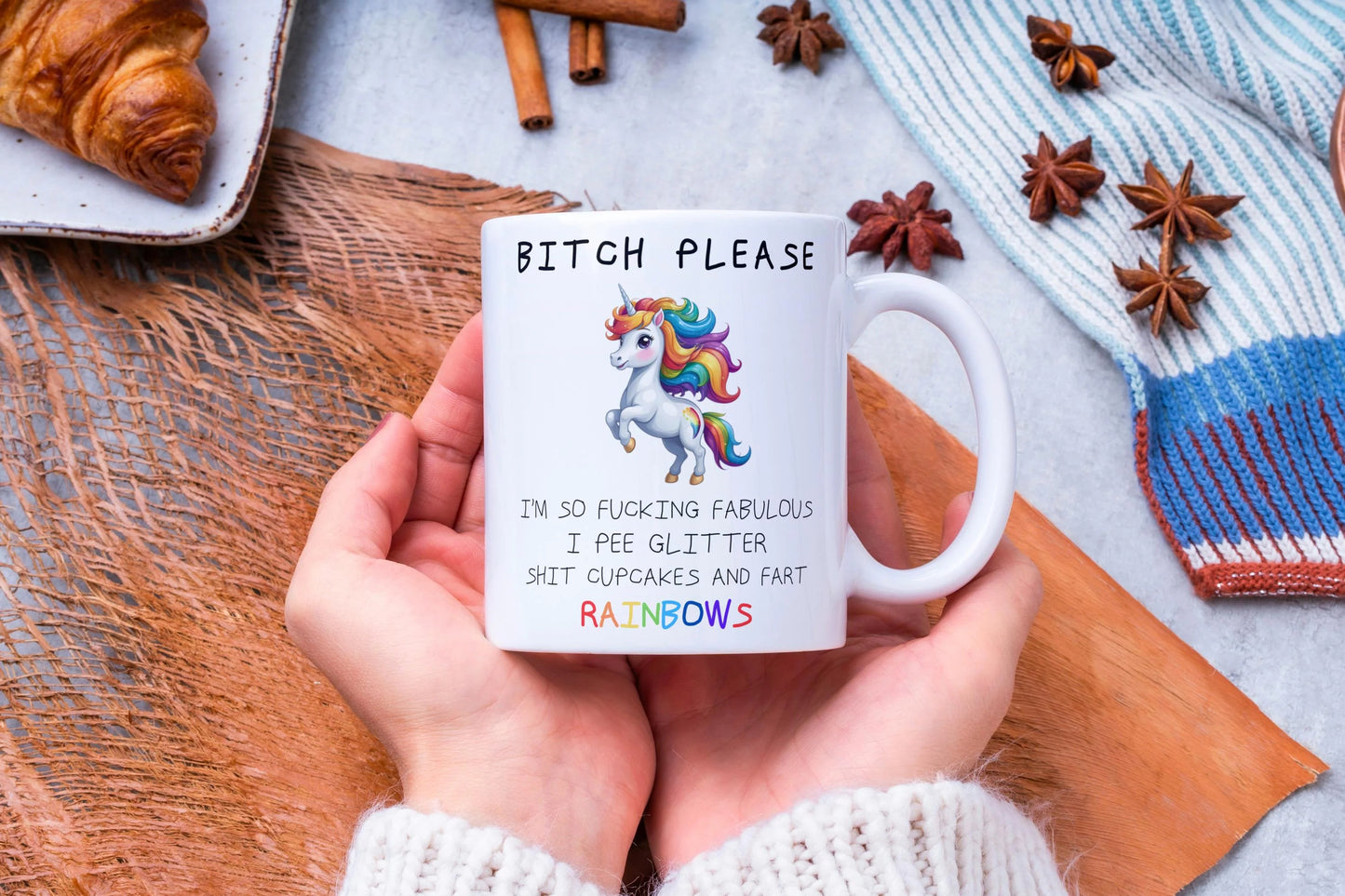 Person holding a mug with a colorful unicorn design and text, surrounded by cinnamon sticks and a croissant on a wooden surface.