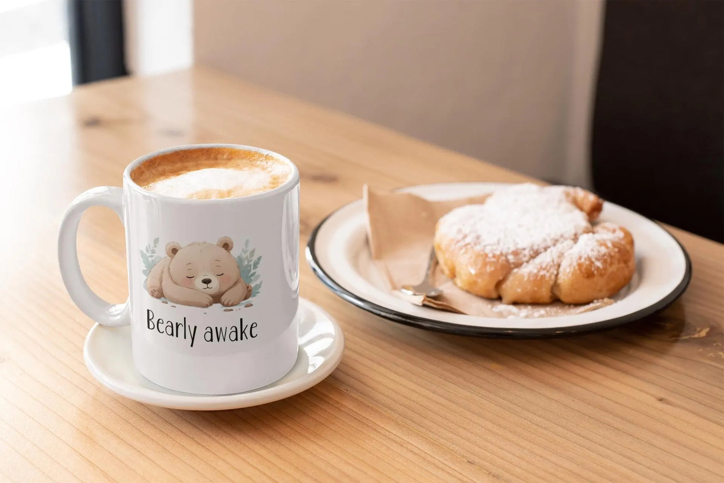 Cup of coffee with a bear design labeled 'Bearly awake' on a saucer, next to a plate with a pastry on a wooden table.