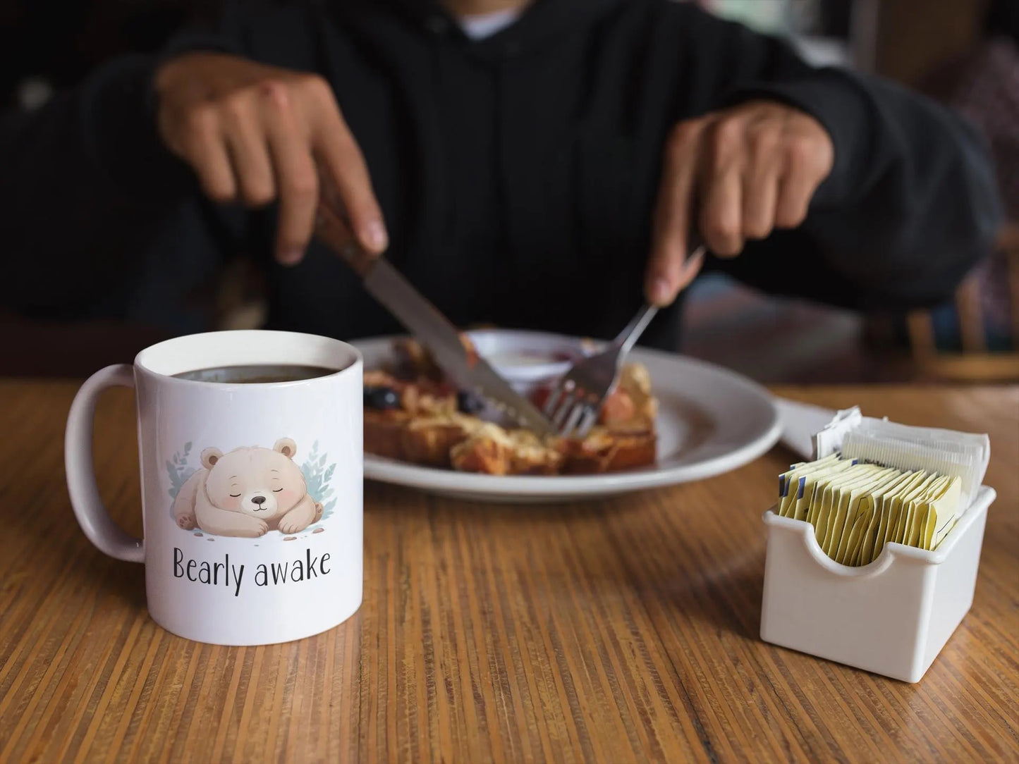 Person eating breakfast with a mug labeled 'Bearly awake' on a table.