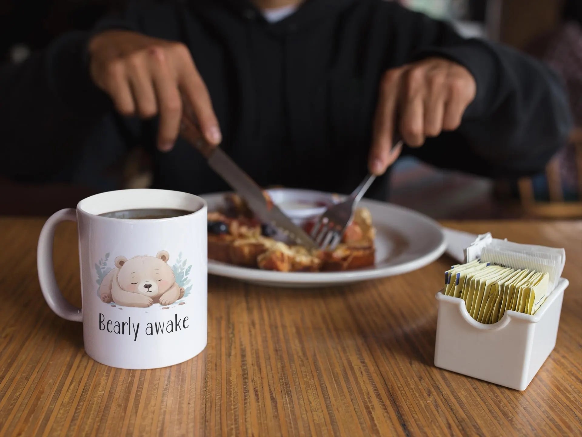 Person eating breakfast with a mug labeled 'Bearly awake' on a table.