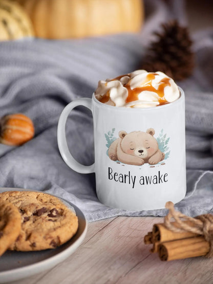 White mug with a bear design and 'Bearly awake' text, filled with hot chocolate and cookies on a wooden surface.
